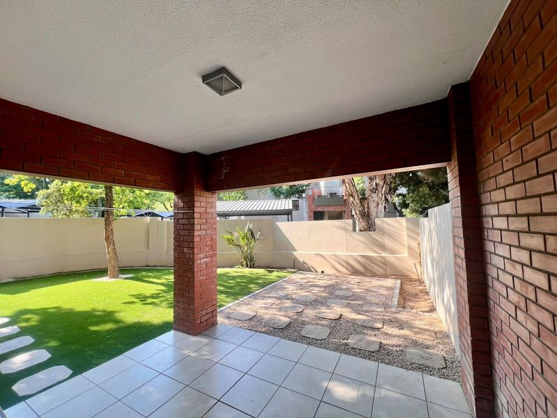 A covered patio featuring exposed brick pillars and tiled flooring, overlooking a private garden area with artificial lawn and stone paving.