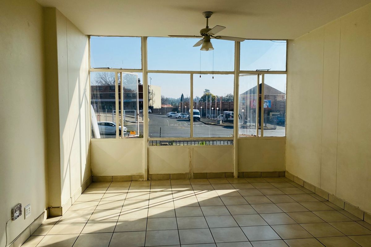 A bright, empty living area featuring tiled flooring, a ceiling fan, and large windows providing natural light and street views.