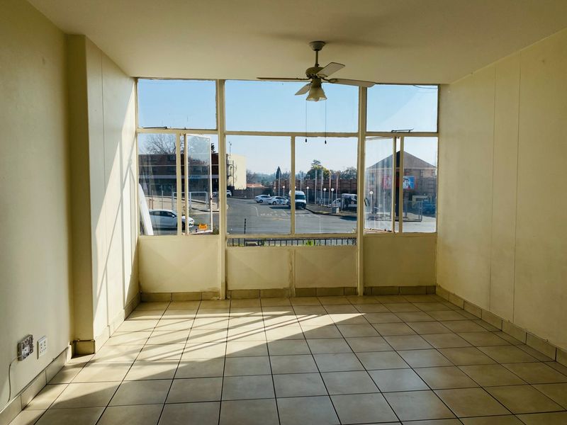 A bright, empty living area featuring tiled flooring, a ceiling fan, and large windows providing natural light and street views.