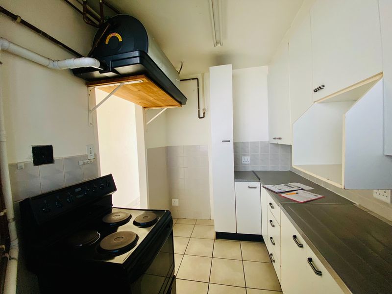 A functional kitchen featuring white cabinetry, dark laminate countertops, tiled flooring, and an electric stove, with exposed plumbing and geyser visible.