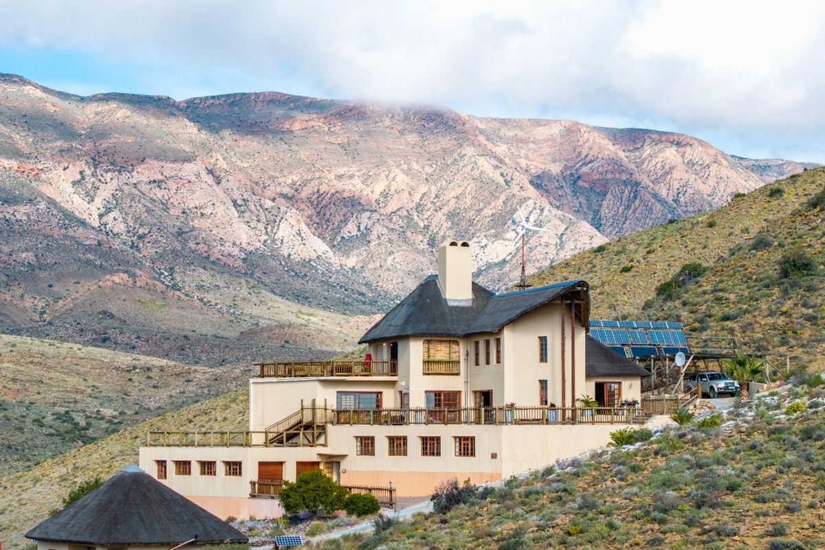 A multi-level farm residence featuring a thatched roof, expansive wooden balconies, integrated solar panels, and a wind turbine, set against a scenic mountain backdrop.