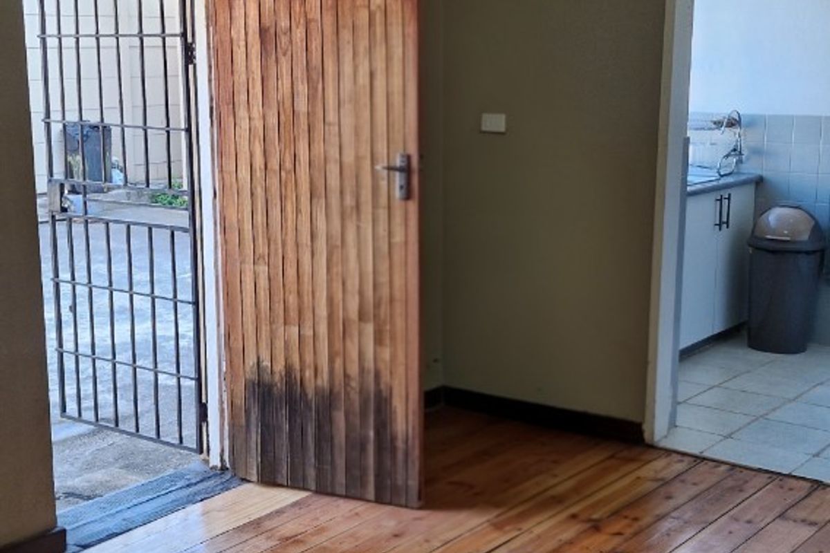 An interior view of a room with polished wooden flooring, featuring a wooden door leading to an exterior area and a doorway into a kitchen.