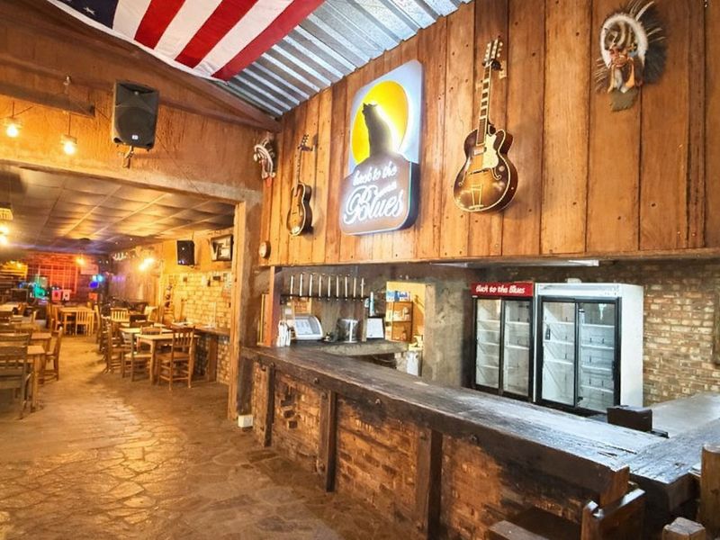 A rustic-style bar area featuring a built-in timber counter, exposed brick walls, stone flooring, and commercial refrigeration units, creating a casual entertainment space.
