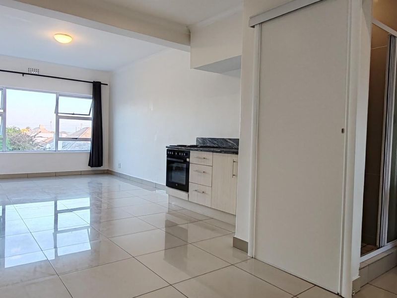 An open-plan apartment layout featuring a kitchen with built-in appliances, tiled flooring, and an adjacent bathroom area with a pedestal basin and shower.