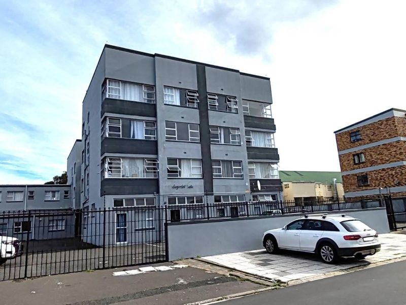 Exterior view of a multi-story apartment building featuring a paved driveway, perimeter fencing, and multiple casement windows.