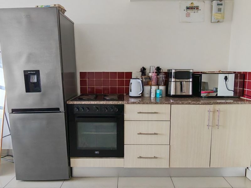 A compact kitchen area featuring light wood-finish cabinetry, granite countertops, a red tiled backsplash, and a built-in oven with an electric hob.