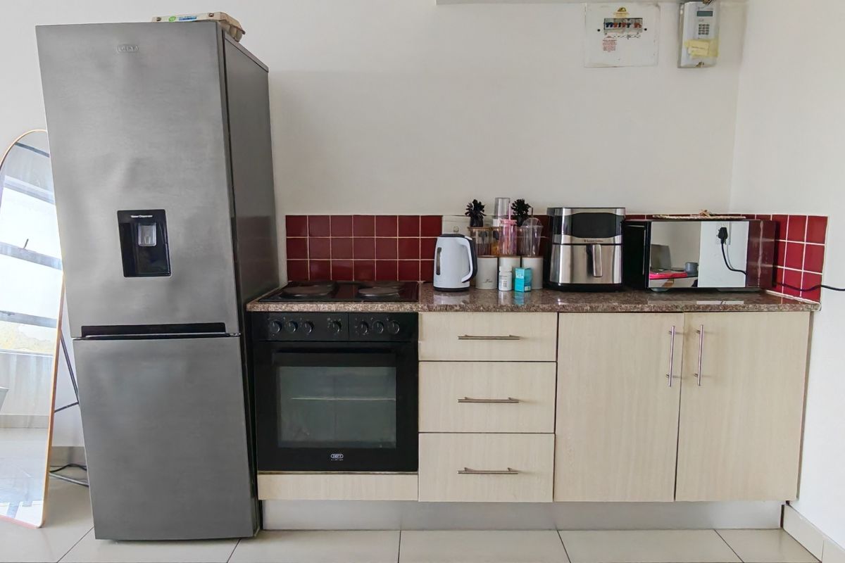 A compact kitchen area featuring light wood-finish cabinetry, granite countertops, a red tiled backsplash, and a built-in oven with an electric hob.