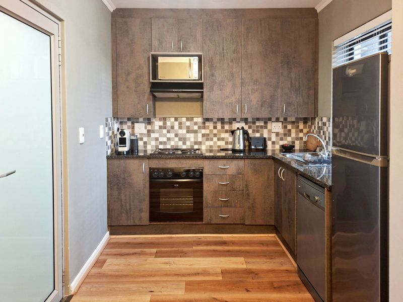 A modern, compact kitchen featuring dark wood-look cabinetry, granite countertops, a mosaic tile backsplash, and integrated appliances including an oven and hob.