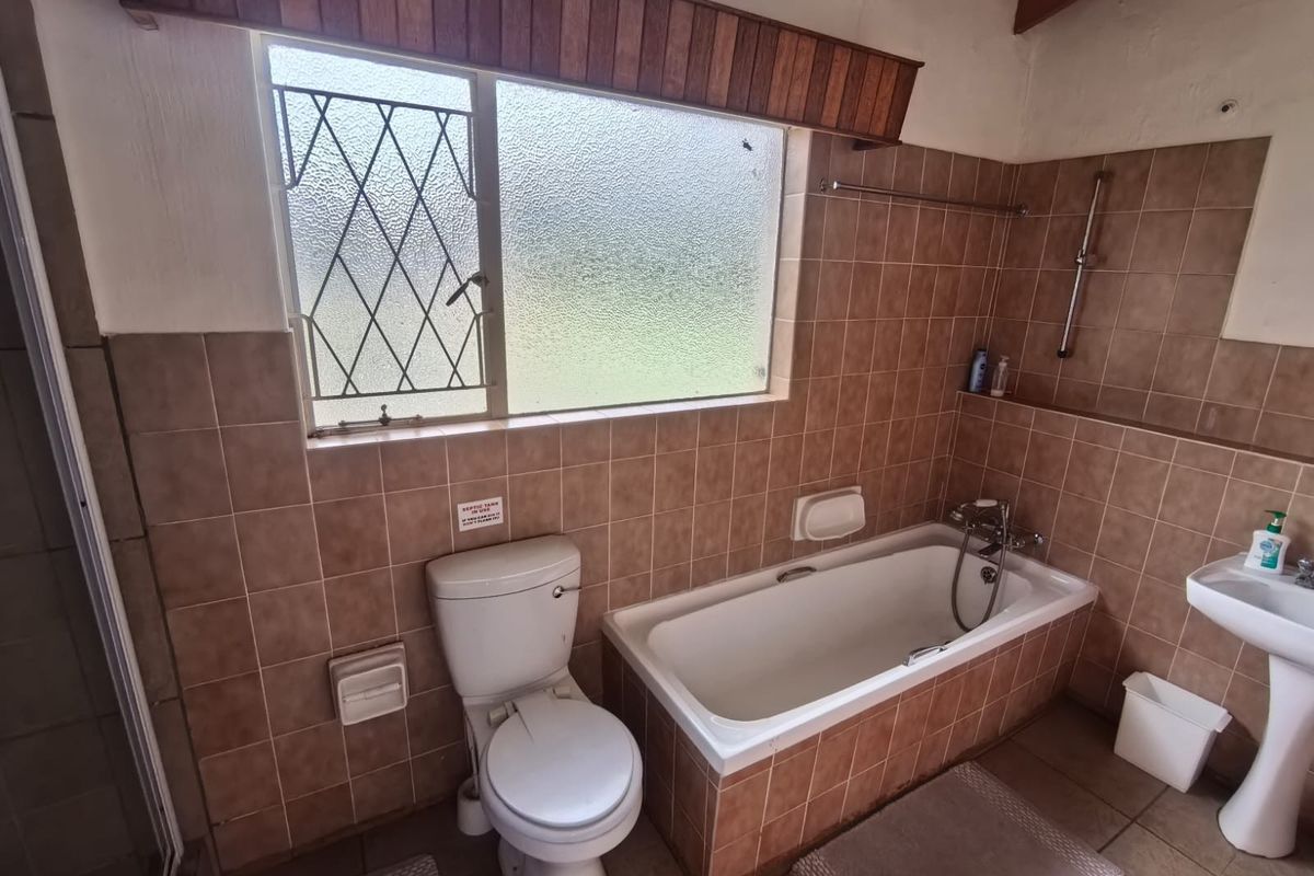 A functional bathroom featuring a built-in bathtub, pedestal basin, toilet, and tiled finishes, complete with security burglar bars on the window.