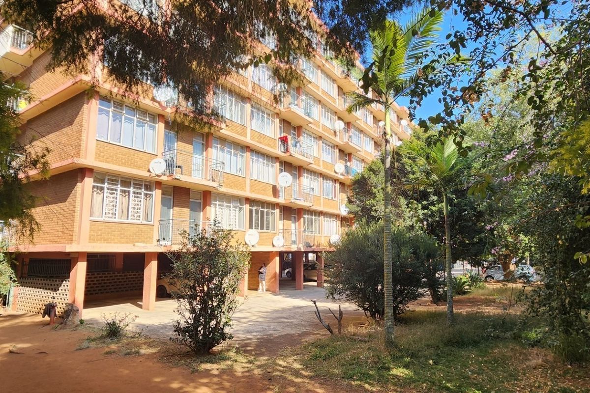 A multi-story apartment building featuring brick facade, large windows, and ground-level covered parking, set within a landscaped garden area.