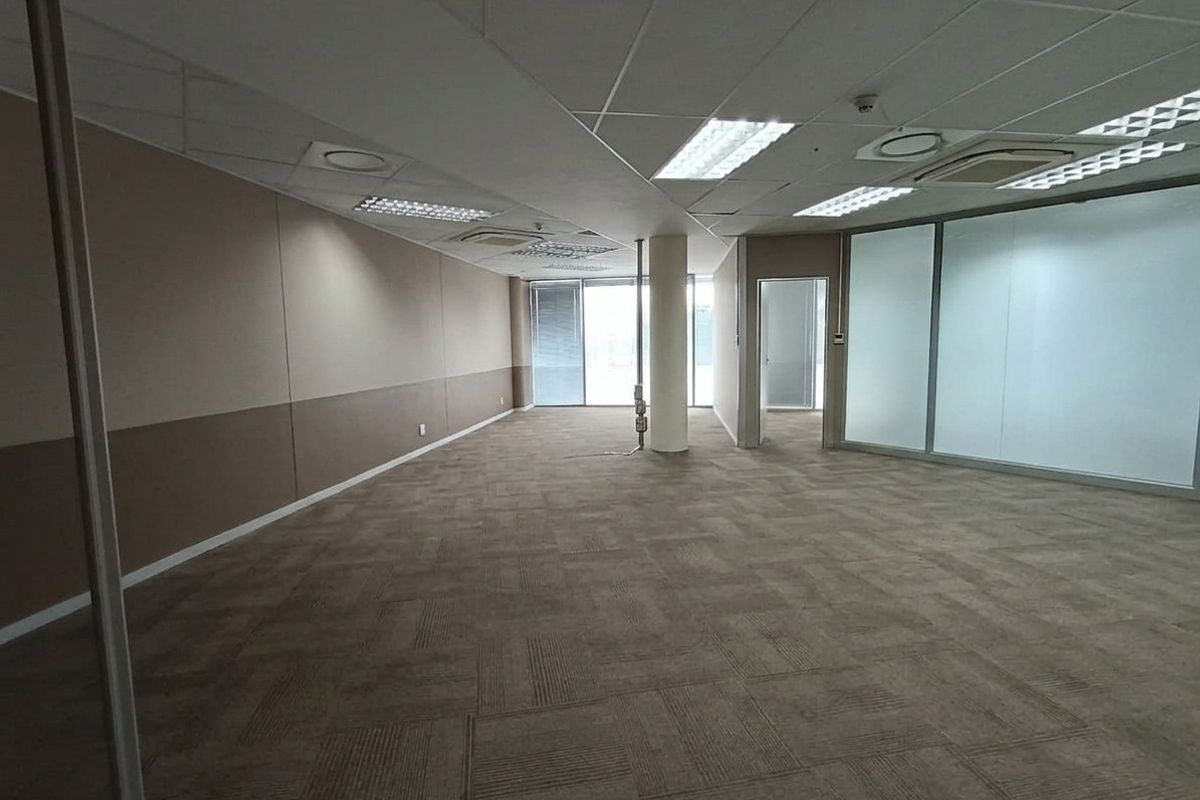 A spacious, empty open-plan office area featuring carpet tiles, suspended ceilings with recessed lighting, and glass partition walls.