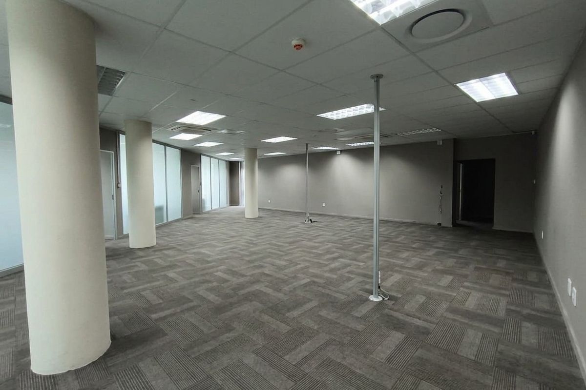 A spacious, open-plan office area featuring neutral carpet tiles, structural columns, and modern suspended ceilings with integrated lighting.
