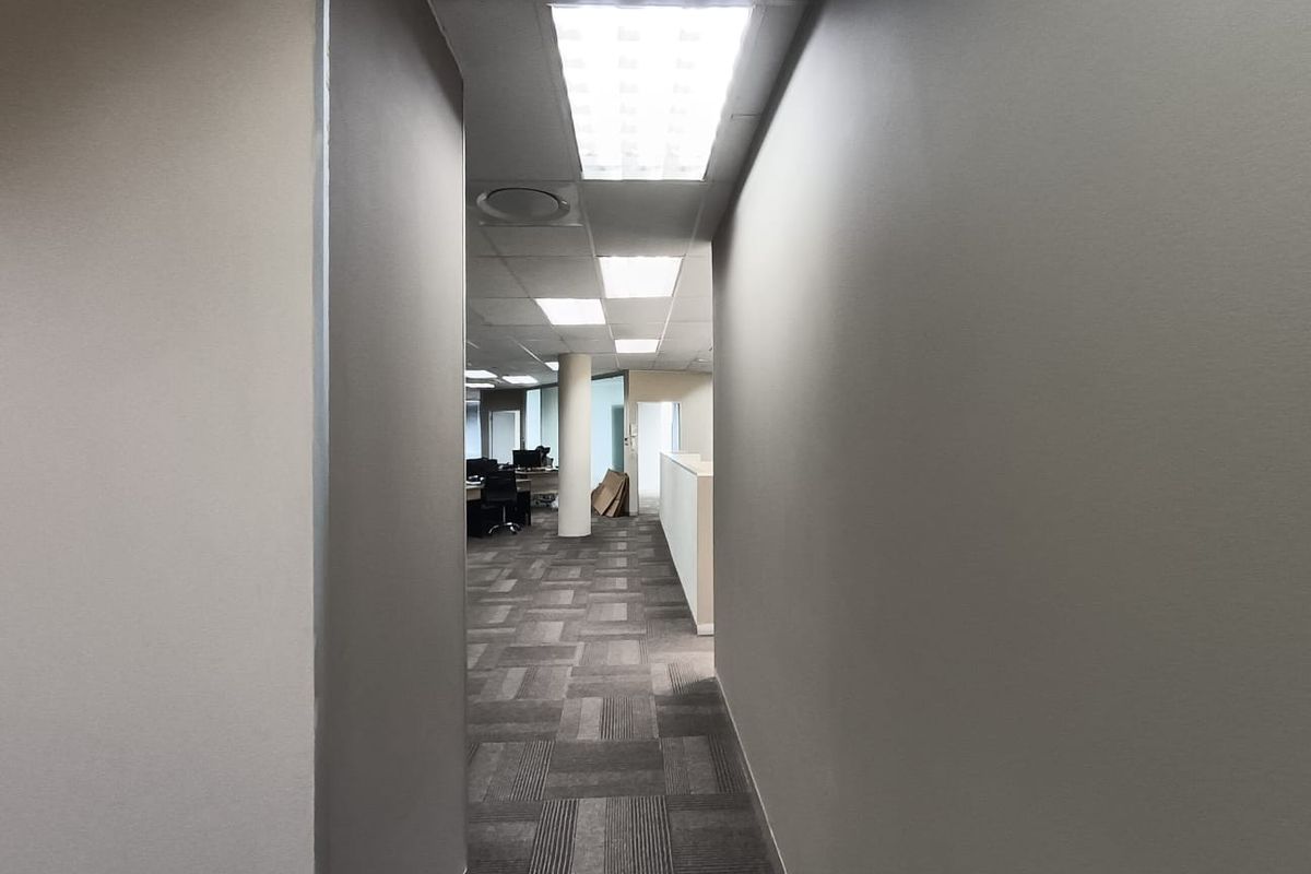 A view through a hallway leading into an open-plan office space featuring suspended ceilings, recessed lighting, and carpet tile flooring.