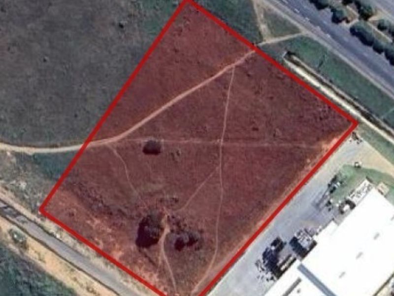 An aerial view of a vacant agricultural plot outlined in red, showing natural terrain and proximity to existing road infrastructure and commercial buildings.