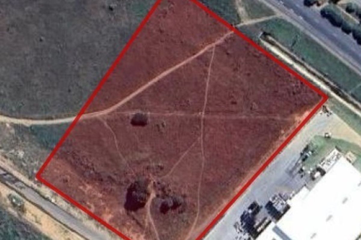 An aerial view of a vacant agricultural plot outlined in red, showing natural terrain and proximity to existing road infrastructure and commercial buildings.