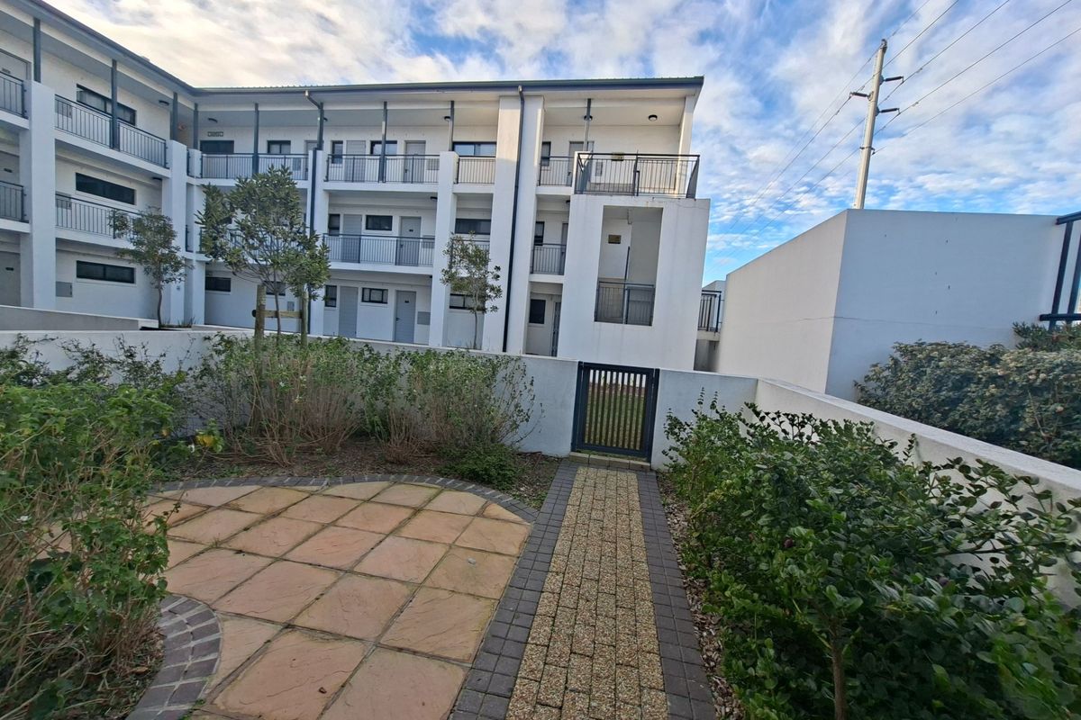 Exterior view of an apartment complex featuring a paved walkway, landscaped garden area, and multi-level balconies with metal railings.