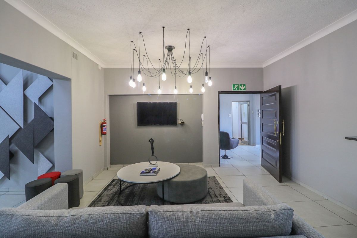 A modern office lounge area featuring tiled flooring, a feature wall with a mounted television, and a contemporary multi-pendant light fixture.