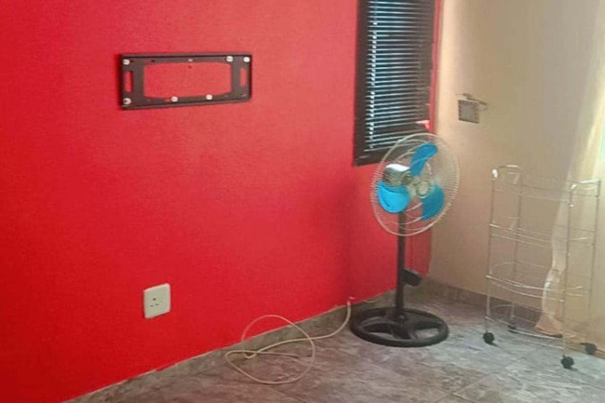 A living room area featuring tiled flooring, a bold red accent wall with a mounted television bracket, and window blinds.