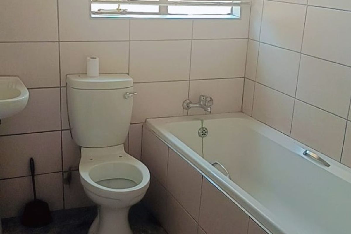A functional bathroom featuring a standard bathtub, toilet, and wall-mounted basin with full-height wall tiling and a window for natural light.