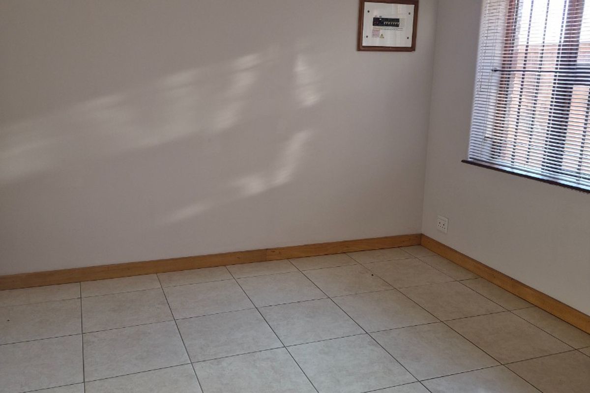 A bright, empty bedroom featuring neutral tiled flooring, wooden skirting boards, and a window fitted with horizontal blinds and security bars.