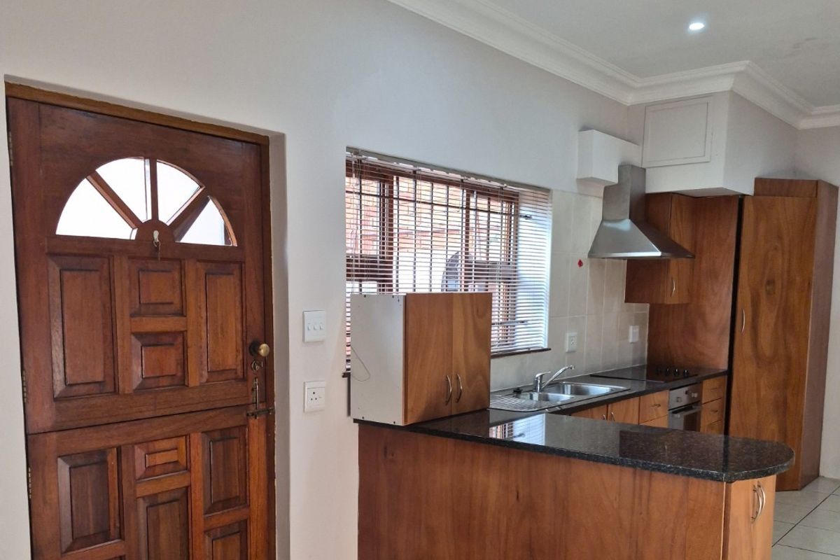 A functional kitchen featuring wooden cabinetry, granite countertops, a stainless steel extractor fan, and a tiled floor, adjacent to a solid wood entrance door.
