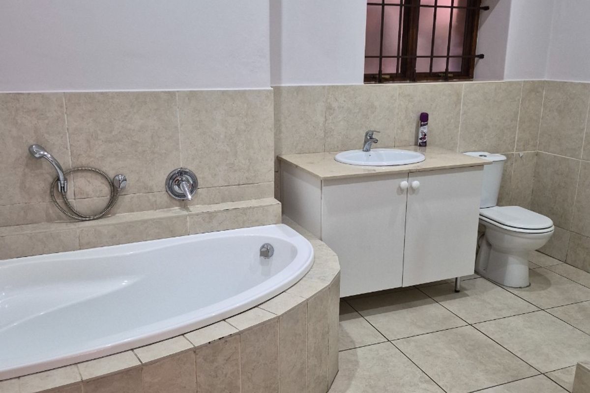 A functional bathroom featuring a built-in tiled bathtub, vanity cabinet with integrated basin, and a toilet, finished with neutral floor and wall tiling.