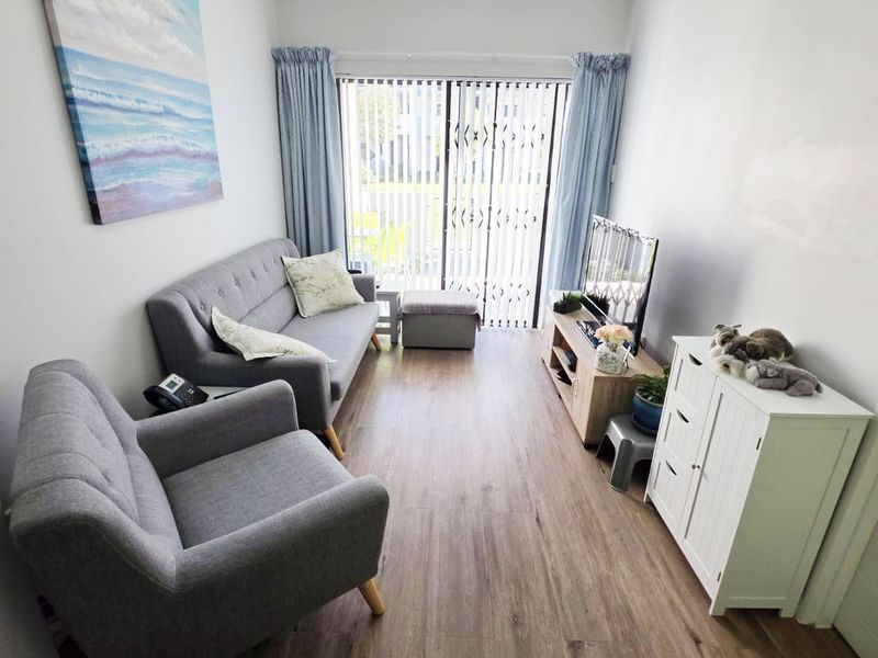 A compact living room featuring laminate flooring, a sliding glass door with security gate, and vertical blinds, offering direct access to the outdoors.