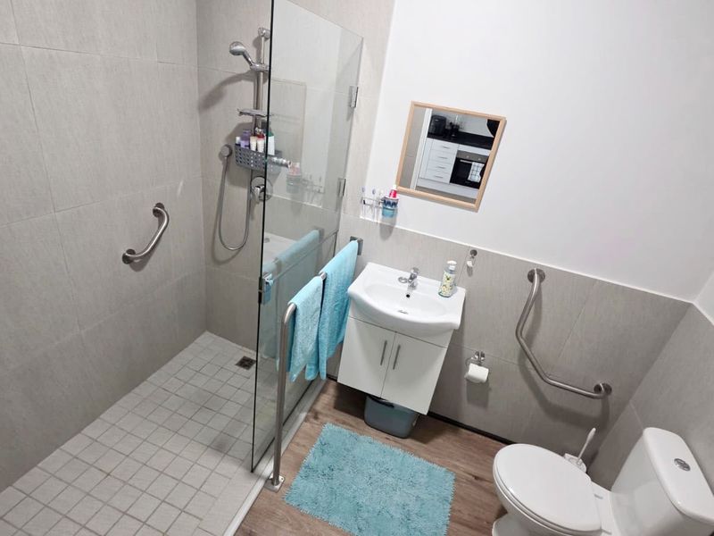 Modern accessible bathroom featuring a walk-in shower with glass screen, vanity basin, toilet, and safety grab rails, finished with neutral wall tiles.