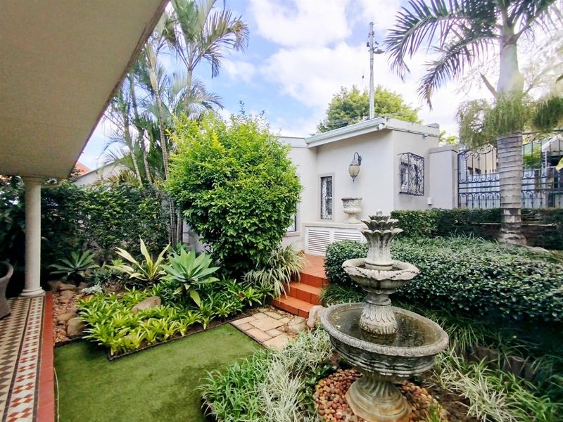 A well-maintained rear garden featuring a stone fountain, landscaped greenery, tiled patio area, and secure wrought iron gate access.