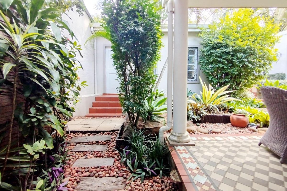 A charming rear courtyard featuring a tiled patio area, stone pathway, and established garden beds leading to an exterior entrance.