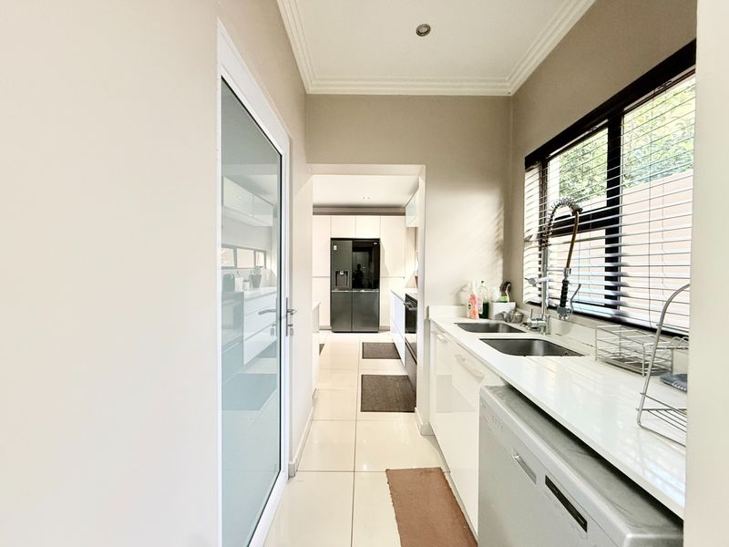 A functional scullery featuring sleek white cabinetry, stone countertops, a double sink with a modern faucet, and tiled flooring, leading into the main kitchen area.