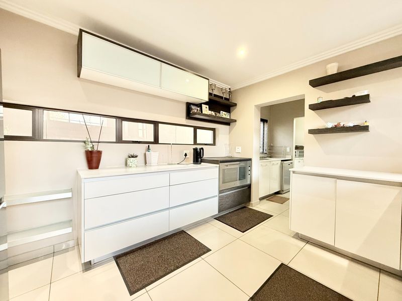 A modern, well-maintained kitchen featuring sleek white cabinetry, built-in oven, floating shelves, and tiled flooring, with an adjacent scullery area.