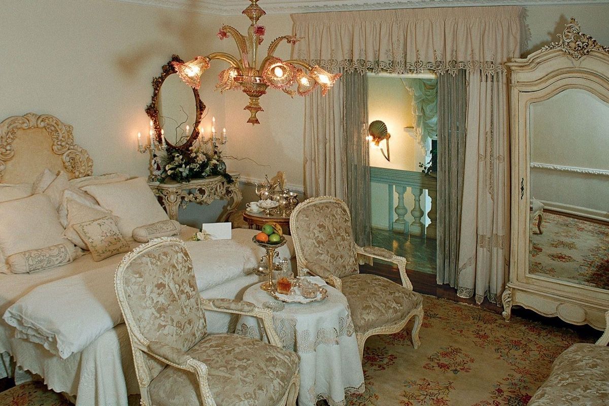 A classic bedroom featuring ornate ceiling cornicing, decorative wall sconces, and a central chandelier, with access to an adjacent hallway area.