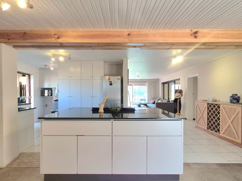 Spacious open-plan kitchen and living area featuring a central island with granite countertops, exposed timber beams, and neutral tiled flooring.