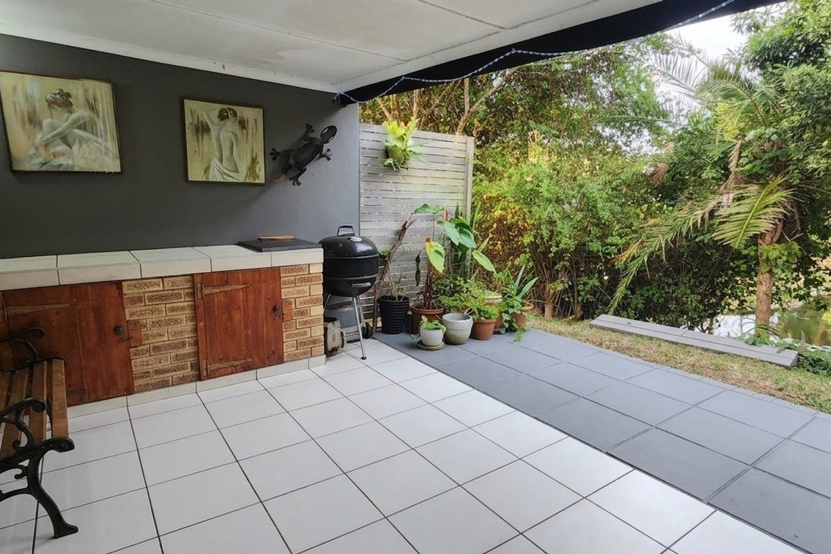 Covered patio area featuring a built-in braai, tiled flooring, and a timber privacy screen, overlooking a lush garden setting.