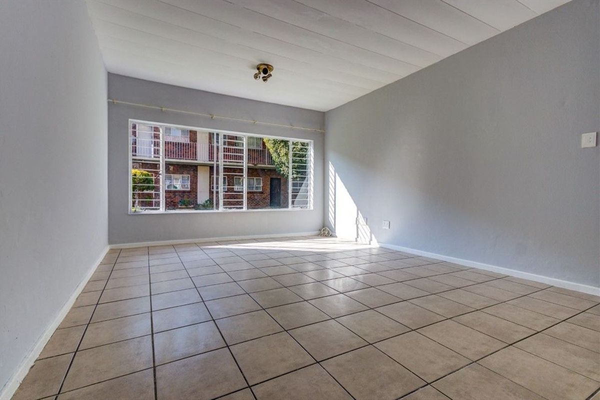 A bright, empty living room featuring neutral tiled flooring, grey walls, and a large window with security bars overlooking the complex.