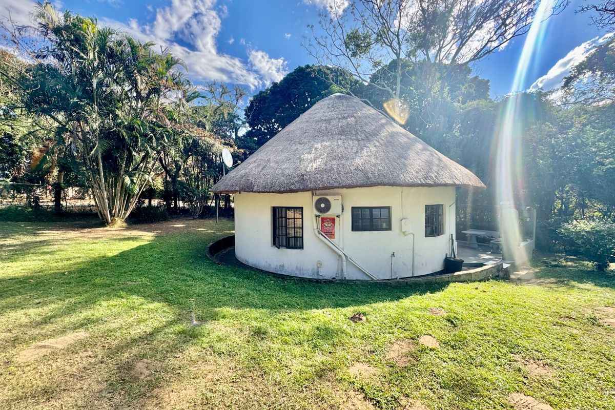 A circular townhouse unit featuring a traditional thatch roof, set within a spacious garden area with mature trees.