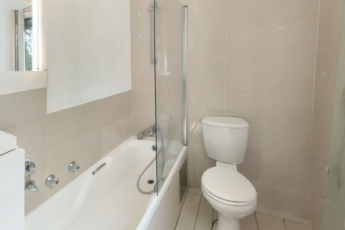 A functional bathroom featuring a bathtub with a glass shower screen, a toilet, and neutral wall and floor tiling.