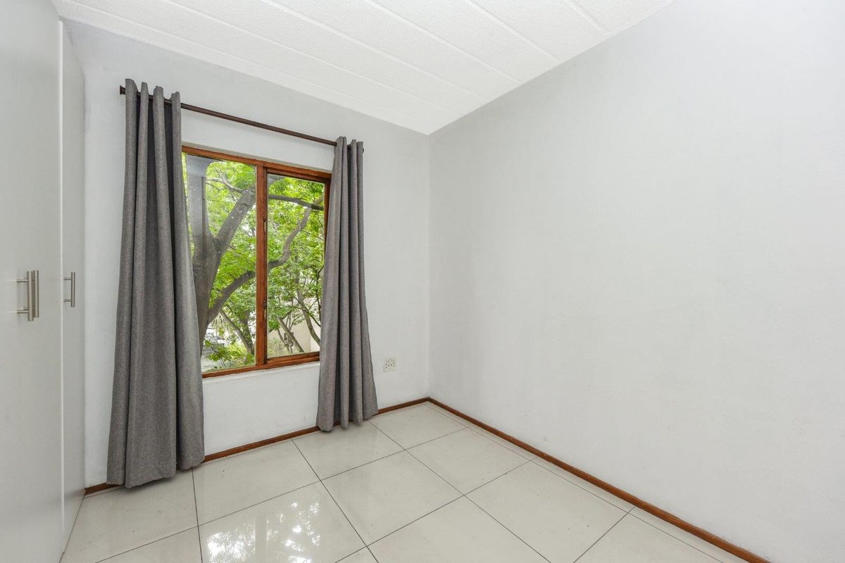 A bright, empty bedroom featuring neutral tiled flooring, a built-in wardrobe, and a wooden-framed window overlooking greenery.