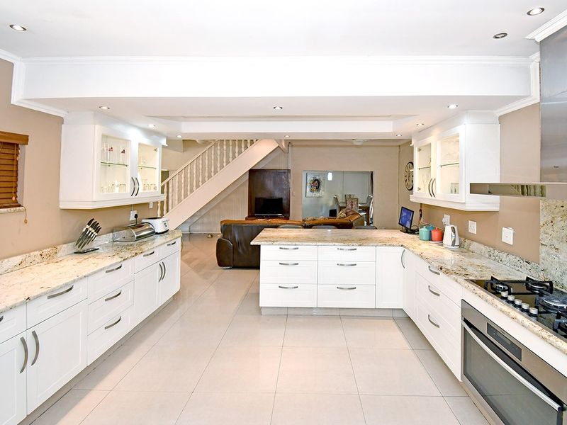 Spacious open-plan kitchen featuring white cabinetry, granite countertops, integrated appliances, and recessed lighting, seamlessly flowing into the living area with a staircase.