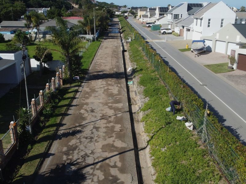 Aerial view of a residential street showing the road infrastructure, drainage channel, and perimeter fencing of the surrounding properties.