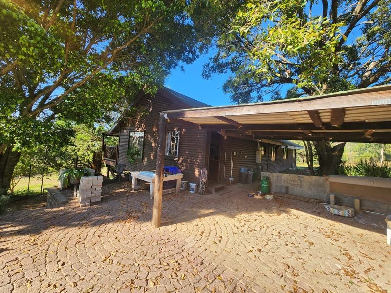 Rear exterior view of a timber log cabin featuring a paved patio area, covered carport, and elevated timber deck nestled among mature trees.