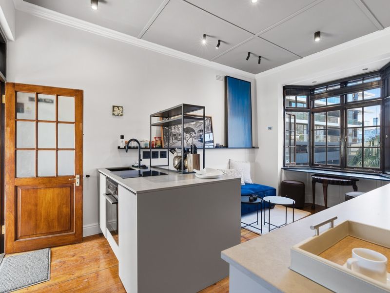 A modern open-plan kitchen and living area featuring a central island, built-in appliances, timber flooring, and a large bay window providing natural light.
