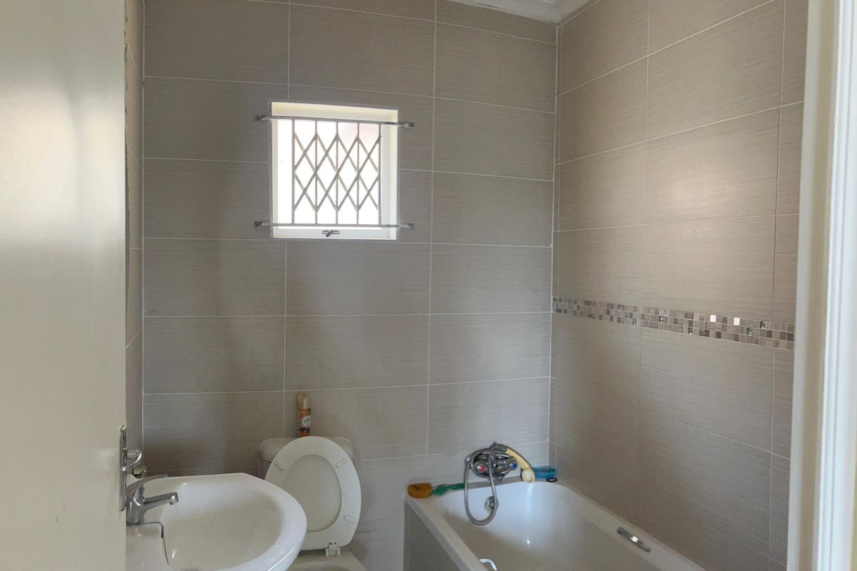 A functional bathroom featuring neutral wall tiling, a standard bathtub with shower mixer, a pedestal basin, and a small window with security bars.