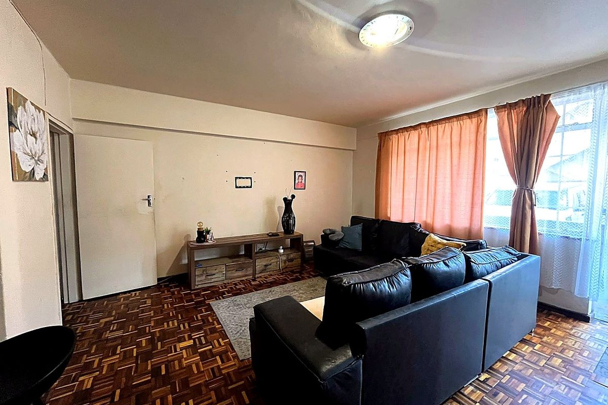 A living room featuring traditional parquet flooring, neutral walls, and large windows with curtains, providing a functional and comfortable living space.
