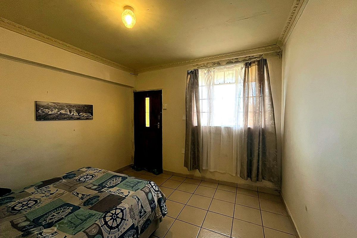 A bedroom featuring tiled flooring, a window with curtains, and a door, showing signs of wear on the ceiling cornice and walls.