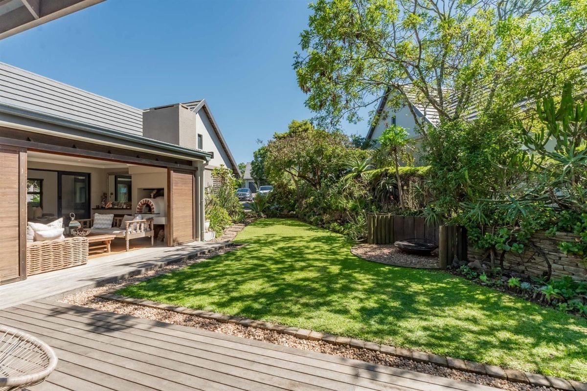 A well-maintained rear garden featuring a timber deck, lush lawn, mature landscaping, and a covered patio area with a built-in pizza oven.