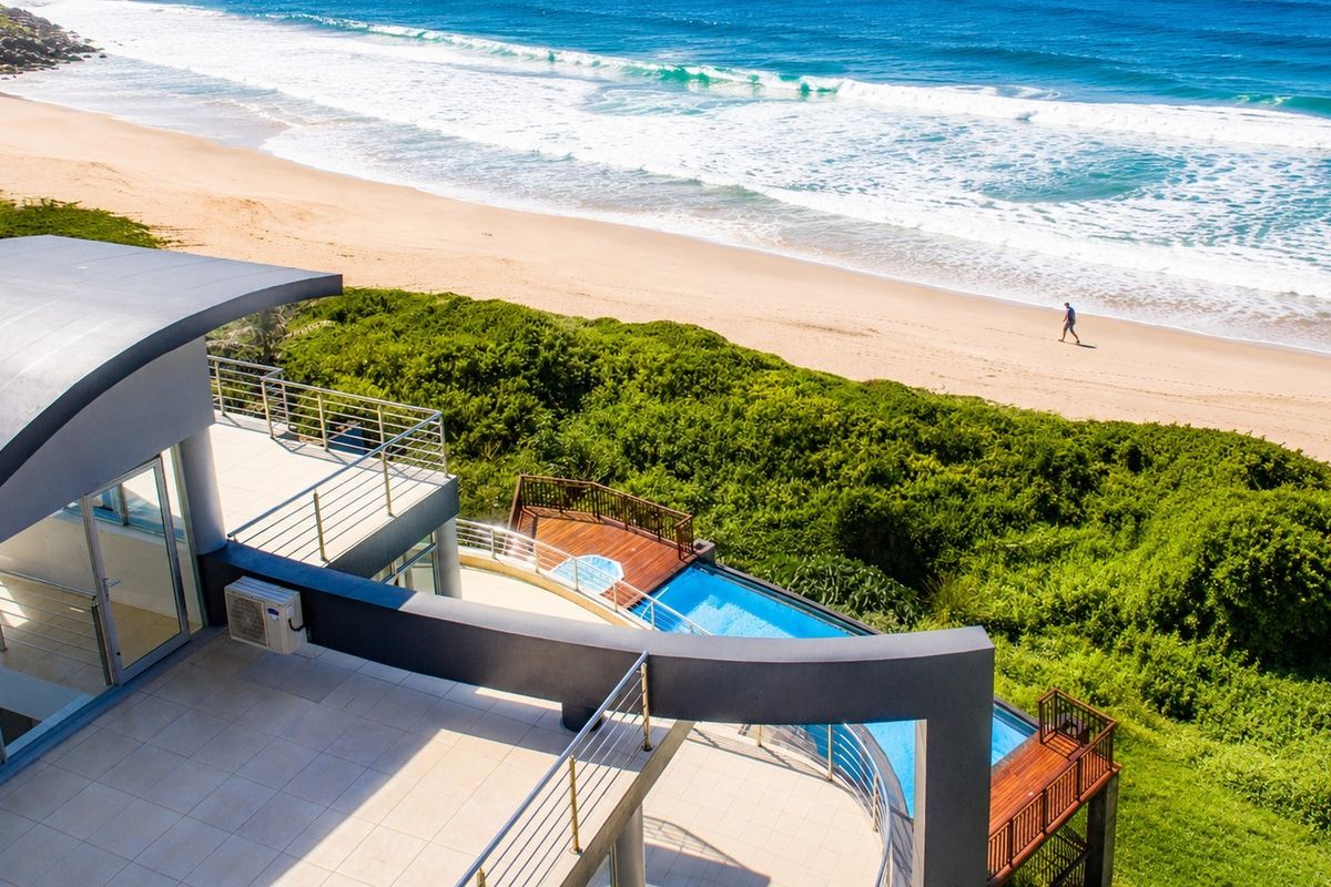 Aerial view of a modern coastal property featuring a swimming pool, timber decking, and expansive tiled patios overlooking the beach.