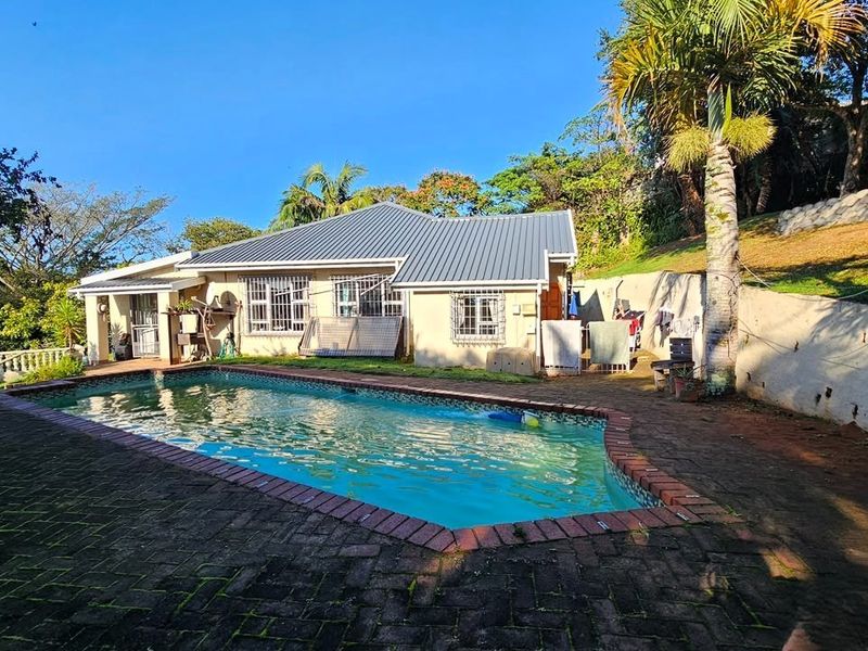 Rear exterior view of the house featuring a swimming pool, brick paved patio area, and a retaining wall set against a landscaped garden.