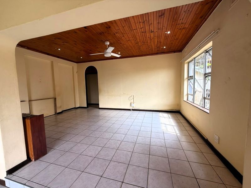 An empty living room featuring tiled flooring, a timber-paneled ceiling with a central fan, and an arched entryway, offering a blank canvas for renovation.